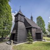 Kościół z ciemnego drewna z niską wieżą. Stoi w otoczeniu drzew, ogrodzony prostym drewnianym płotem.