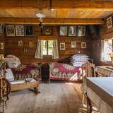 Wnętrze starej drewnianej, chłopskiej chaty, stół, dwa łóżka bogato zasłane kolorowymi kapami, wiele poduszek, dużo obrazków w drewnianych ramach na ścianach, drewniany kołowrotek.