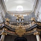 Widziane z dołu kościelne organy na chórze, bogate zdobienia, pozłacane figury. W centralnym punkcie na złotej tablicy napis: IN OMNEM TERRAM EXIVIT SONUS EORUM.